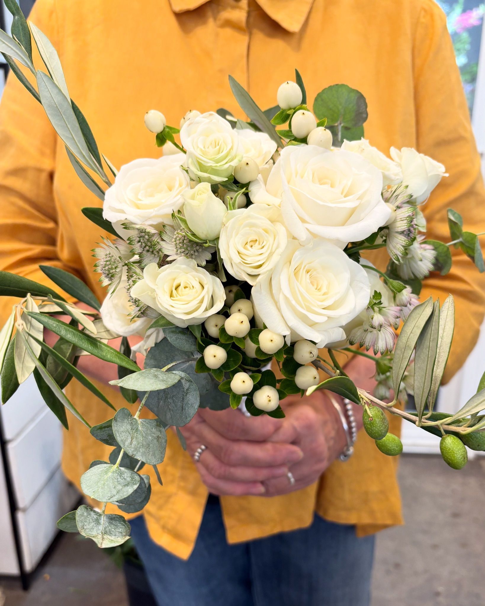 Une personne tenant un bouquet de roses blanches, d'eucalyptus et de verdure devant une chemise jaune moutarde.