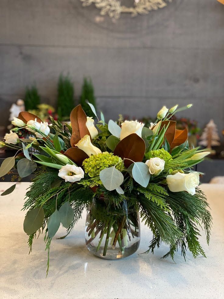 Centre de table floral avec roses blanches, feuillage vert et feuilles cuivrées dans un vase en verre