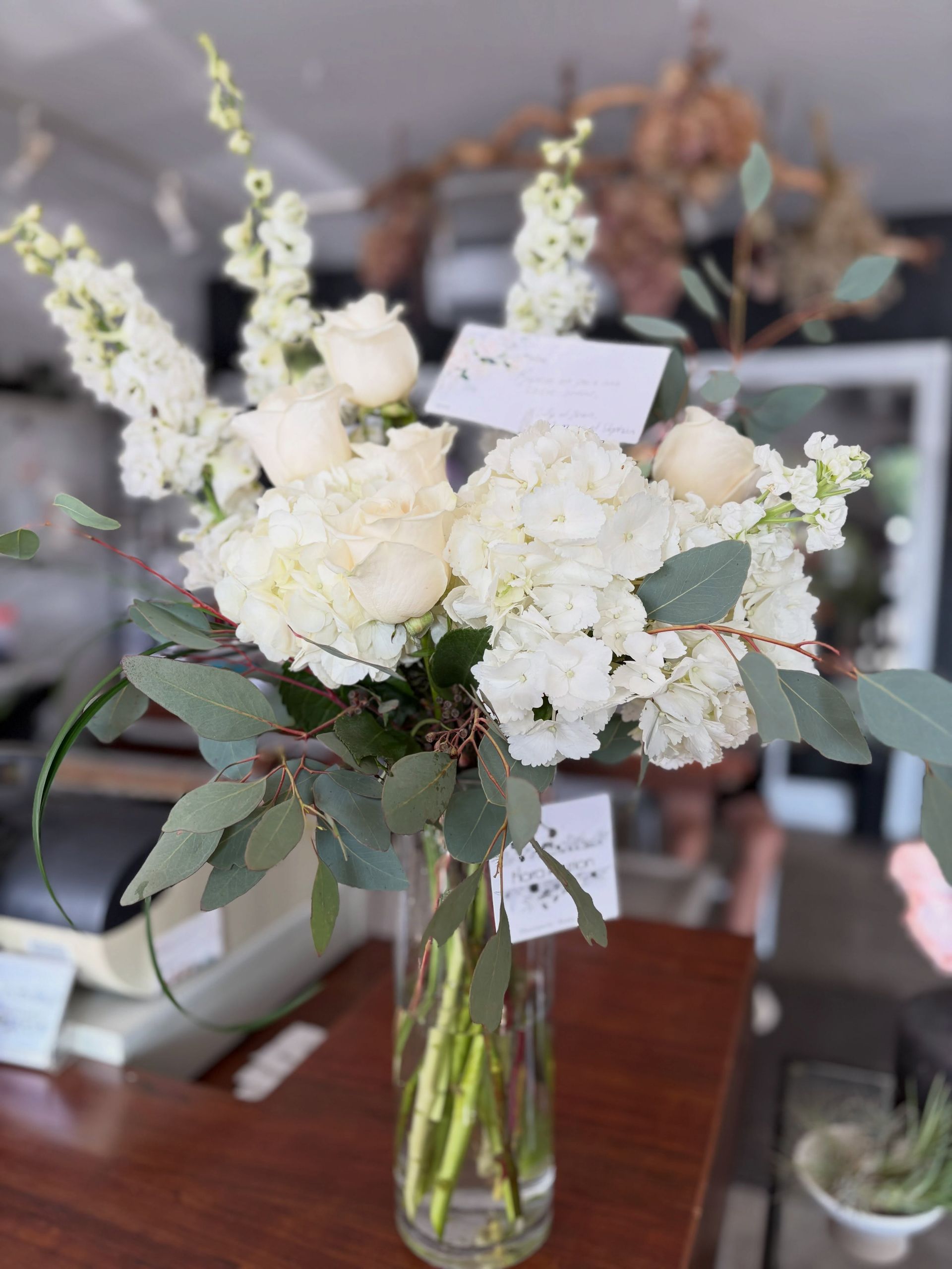 Bouquet de fleurs blanches dans un vase en verre posé sur une table en bois, agrémenté de feuillage vert et d'une petite carte.