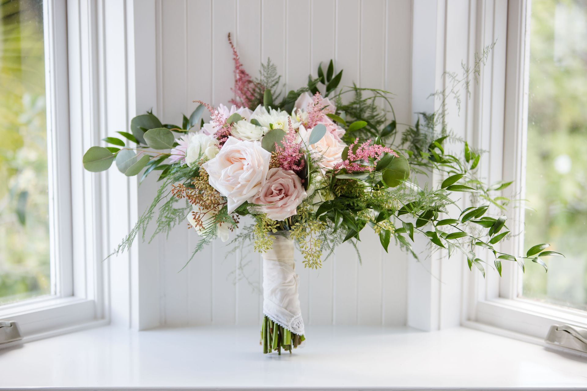 Bouquet de fleurs rose pastel et blanc avec de la verdure dans un vase sur un rebord de fenêtre blanc
