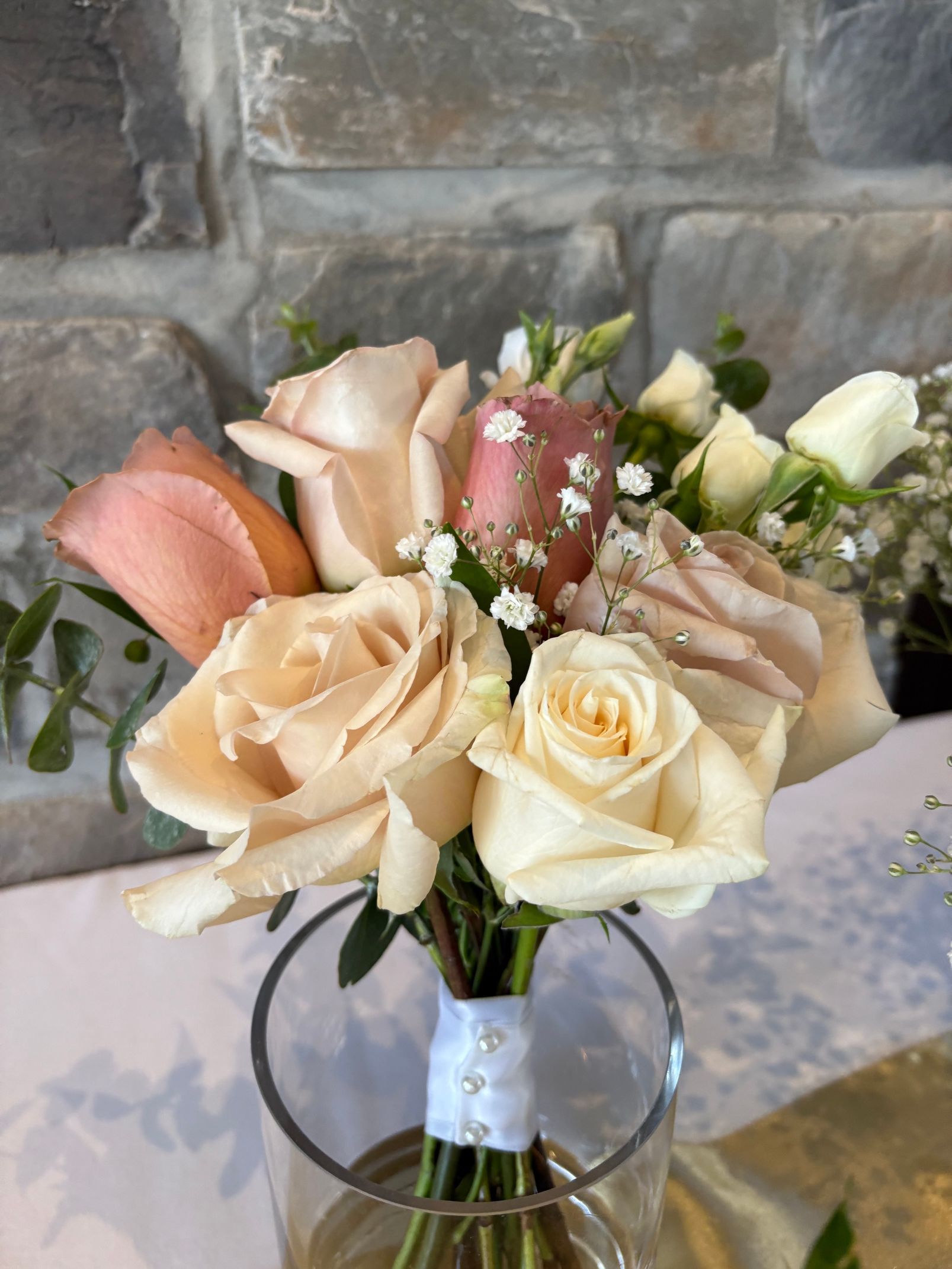 Bouquet de roses crème et rose poudré dans un vase en verre, devant un mur de pierre.