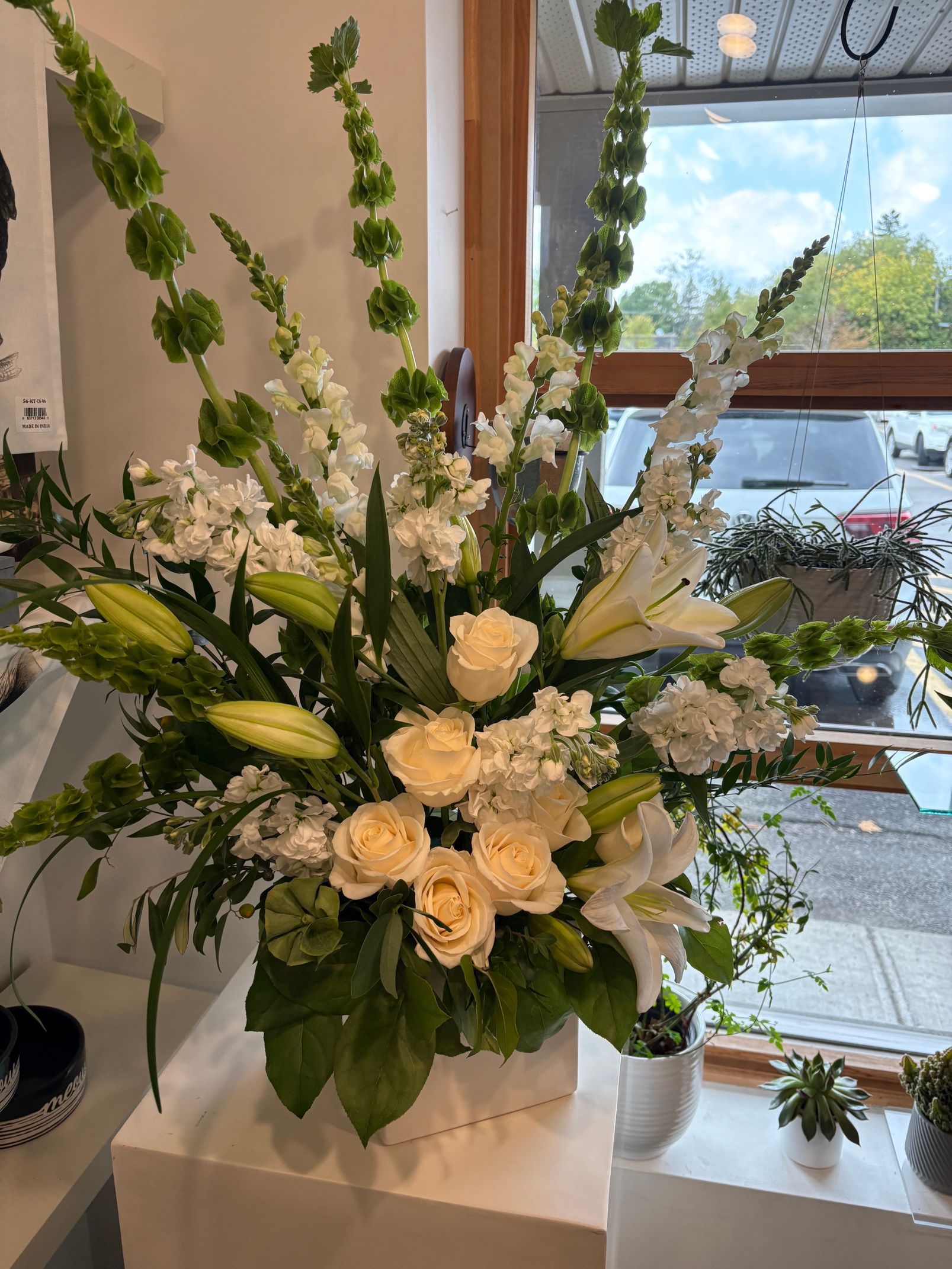 Composition florale blanche dans un vase sur un rebord de fenêtre, avec de la verdure et une vue lumineuse sur une vitrine en arrière-plan.