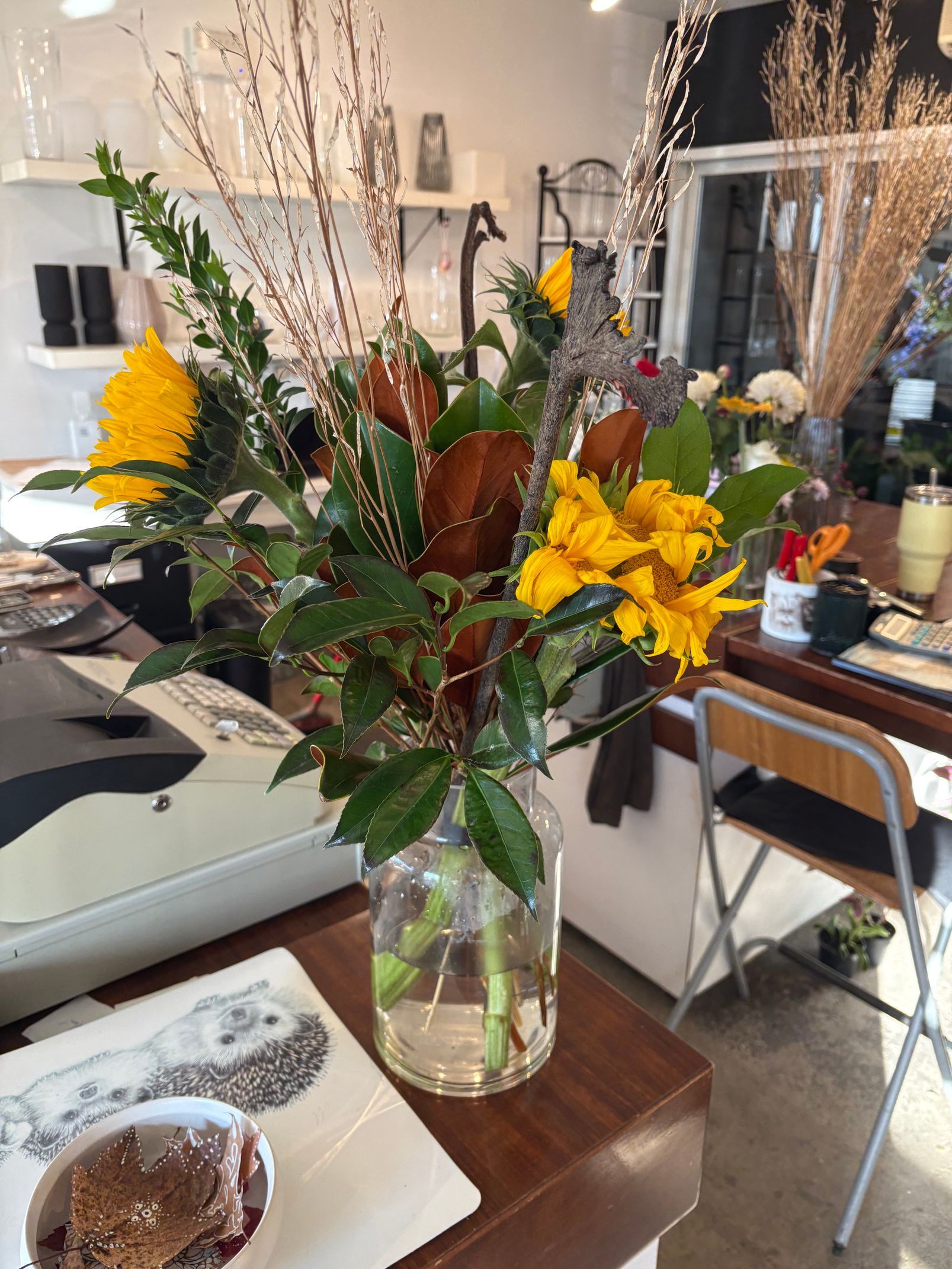 Bouquet de fleurs jaunes et séchées dans un vase en verre, posé sur une table en bois dans un café lumineux.