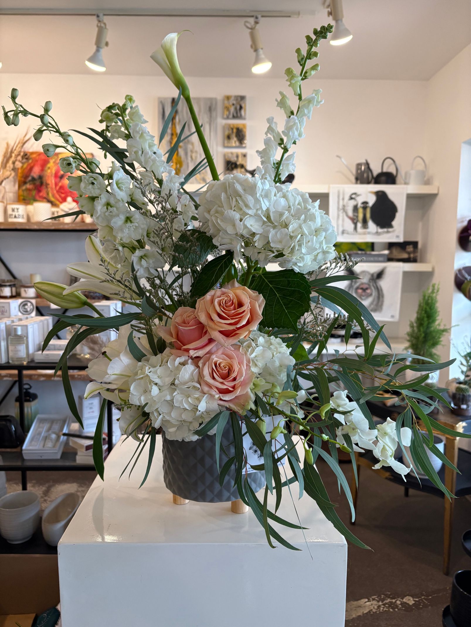 Grand arrangement floral pastel composé de fleurs blanches et roses dans un vase posé sur un piédestal, à l'intérieur d'un magasin.