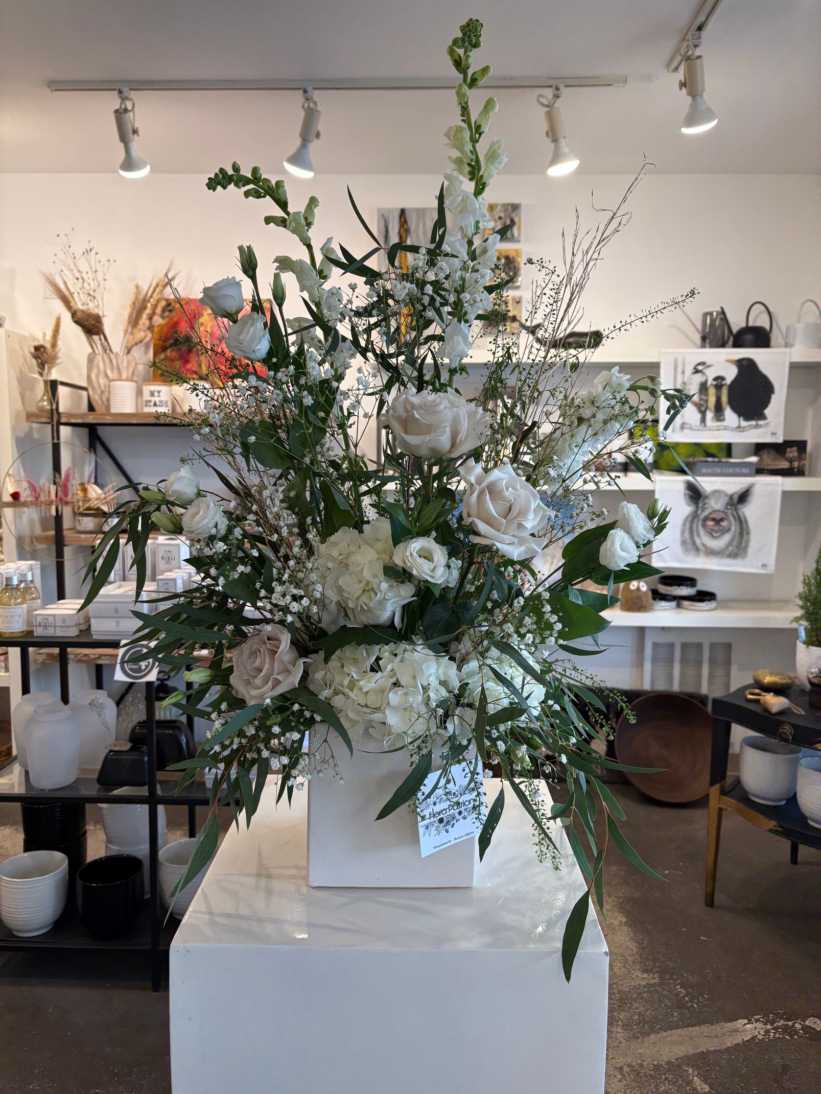 Grand arrangement floral blanc avec de la verdure dans un vase, exposé dans une boutique-atelier lumineuse.