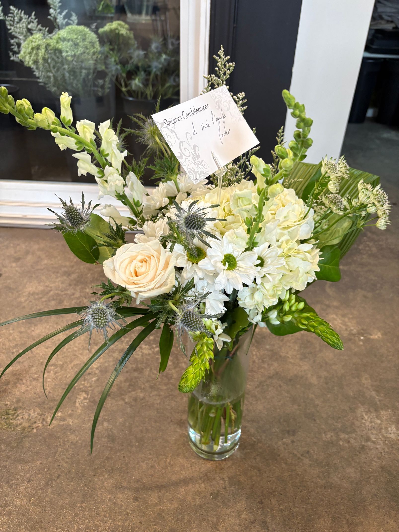 Composition florale blanche et verte dans un vase en verre avec une carte de vœux, présentée sur une table à l'intérieur.
