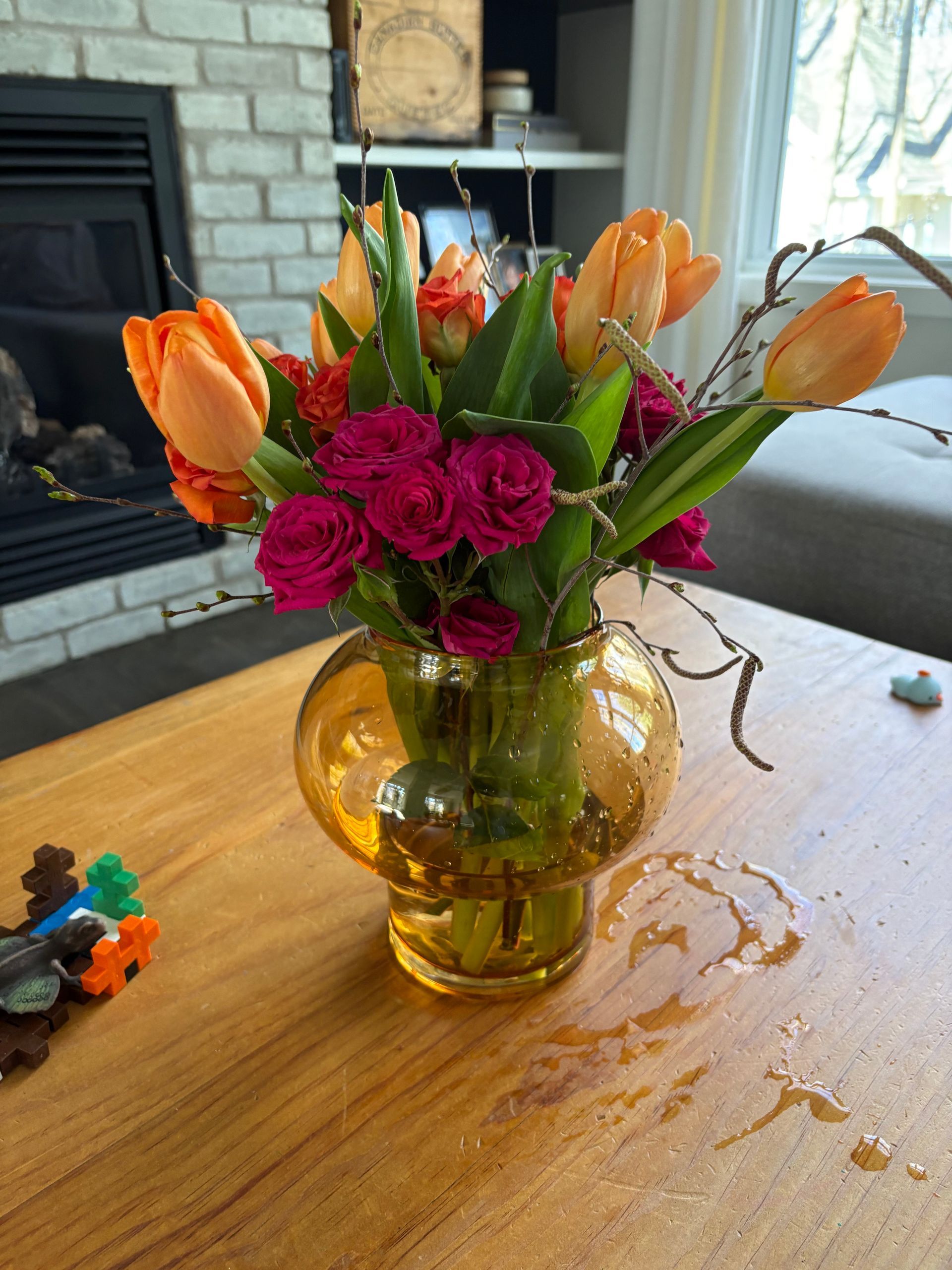 Des tulipes orange et des roses roses dans un vase en verre jaune sur une table en bois près d'une cheminée