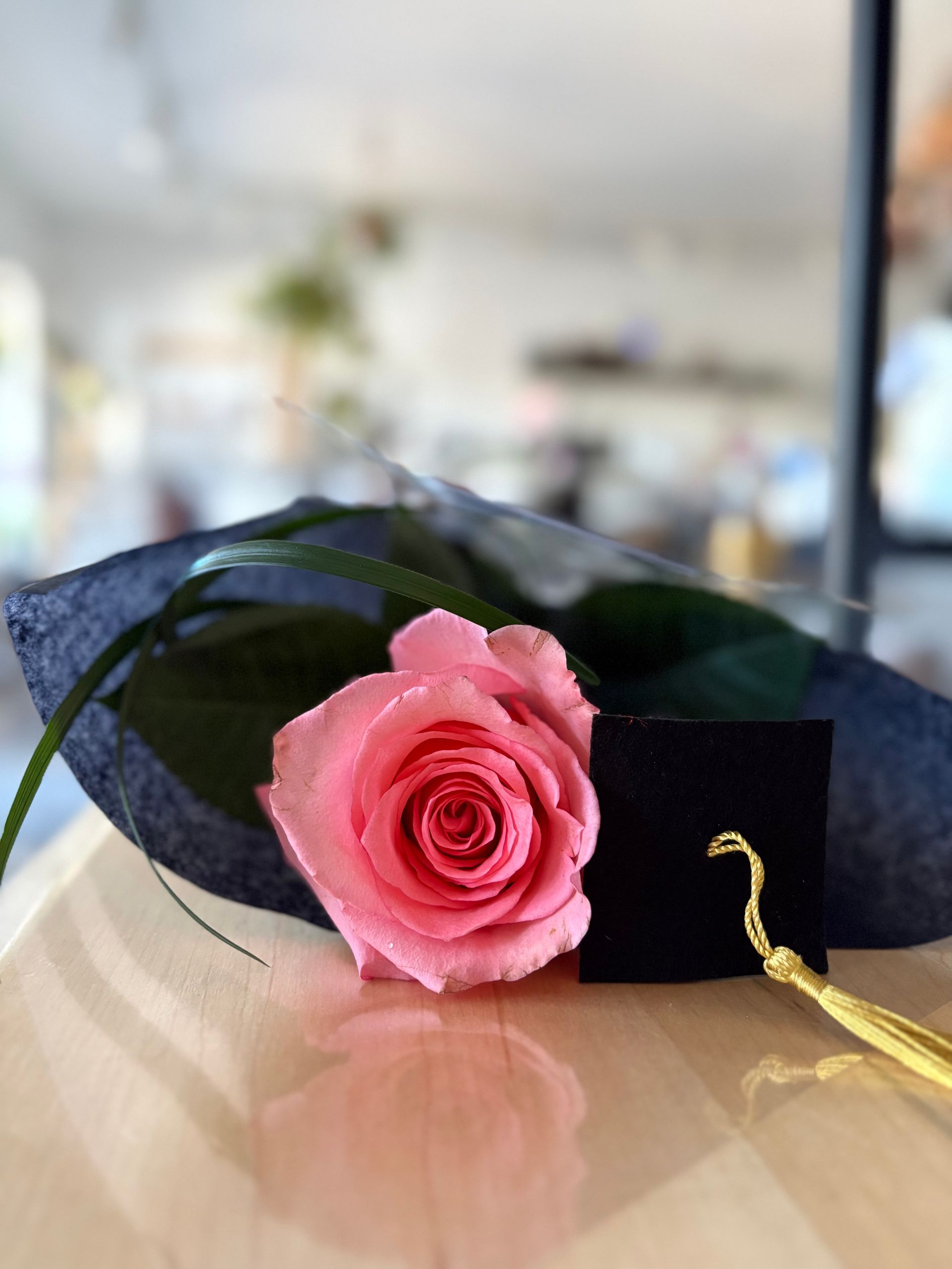 Une rose rose et une toque de diplômé noire sur une table, sur un fond d'intérieur flou.