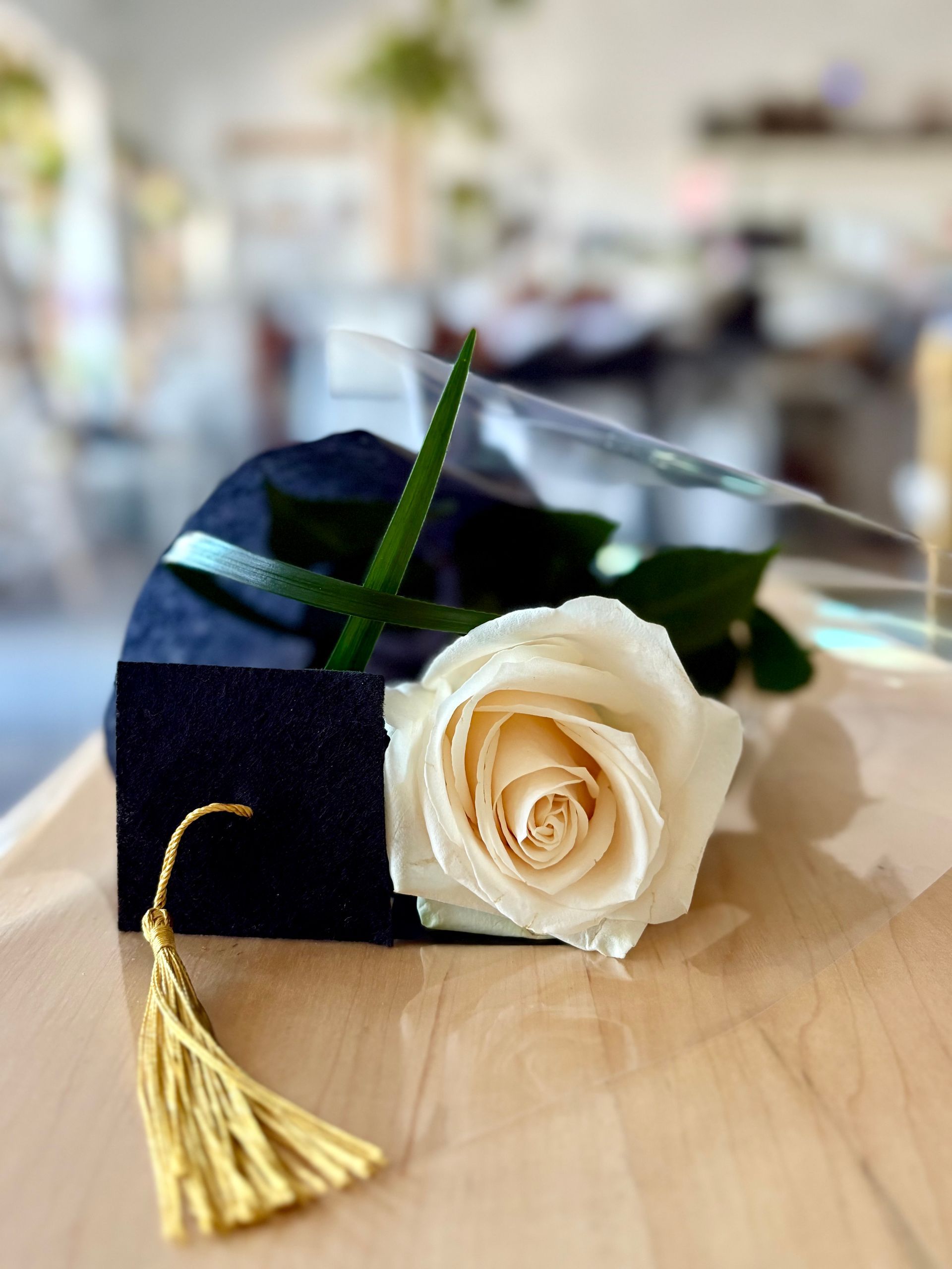 Toque de graduation noire ornée d'une rose blanche et d'un pompon doré posée sur une table, sur fond flou.