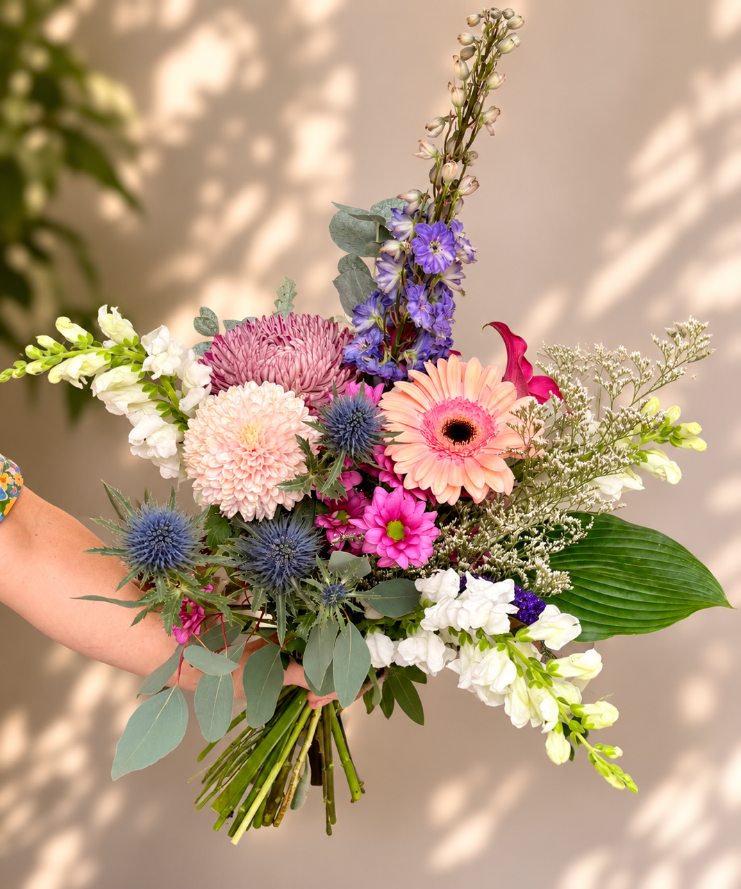 Bouquet de fleurs roses, violettes, blanches et bleues, noué à la main, avec un feuillage vert sur un fond beige clair.