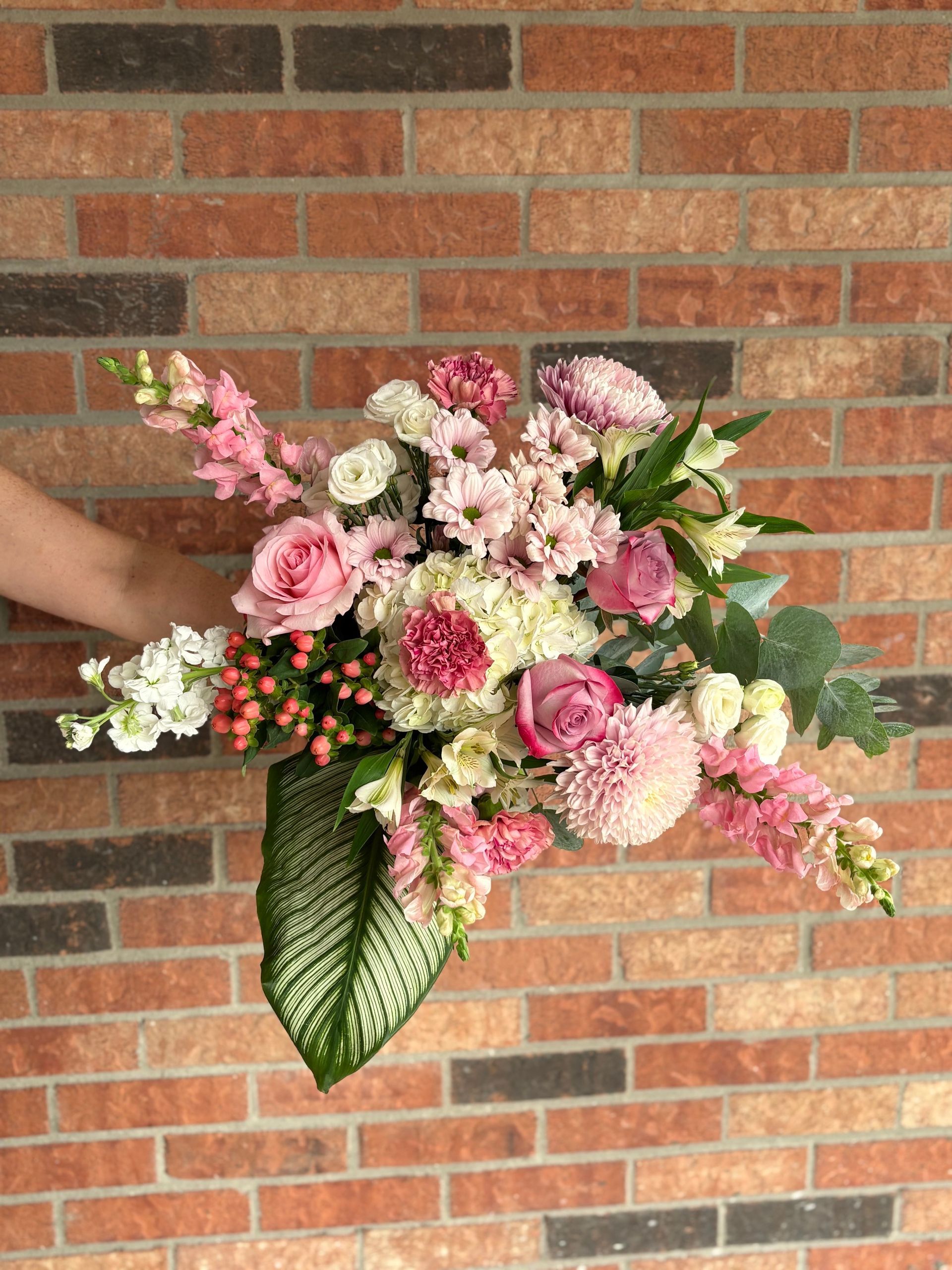 Bouquet de fleurs roses, blanches et vertes porté à la main devant un mur de briques