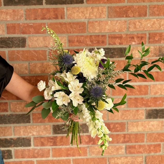 Bouquet de fleurs blanches, jaunes et violettes porté à la main devant un mur de briques