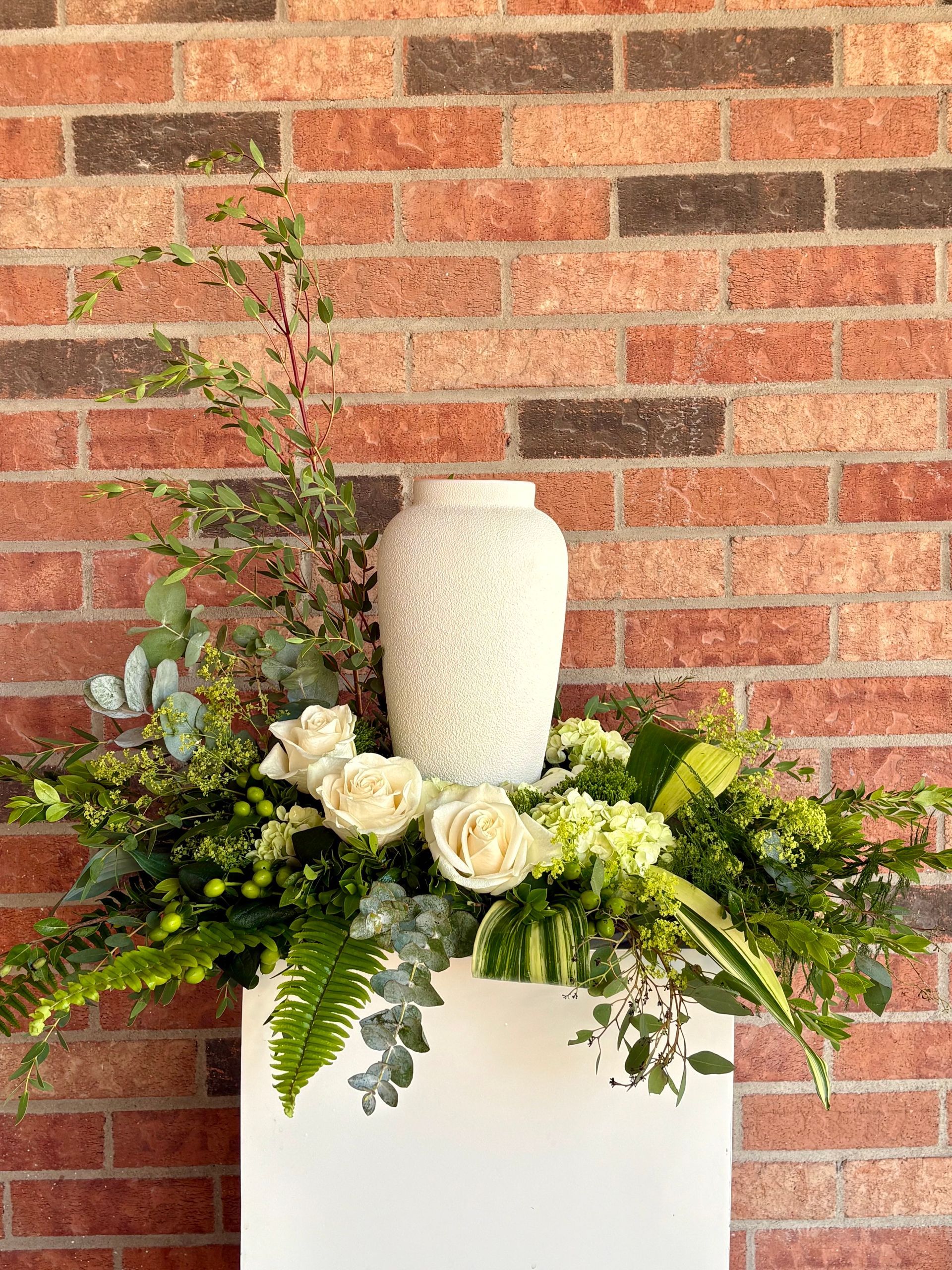 Composition florale blanche avec de la verdure et un vase crème sur un piédestal blanc devant un mur de briques