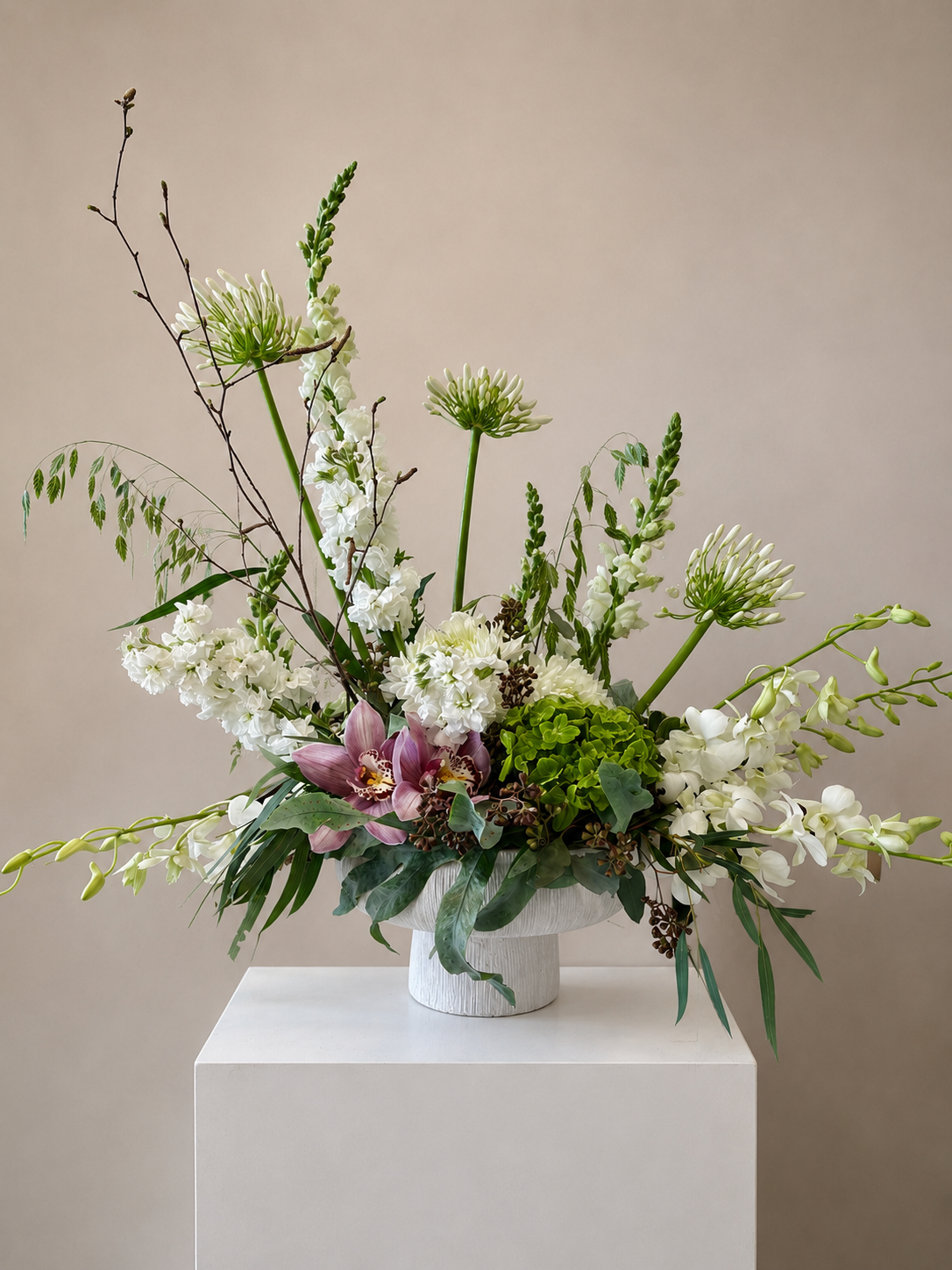 Composition florale blanche avec feuillage vert et touches de rose dans un vase sur un piédestal.