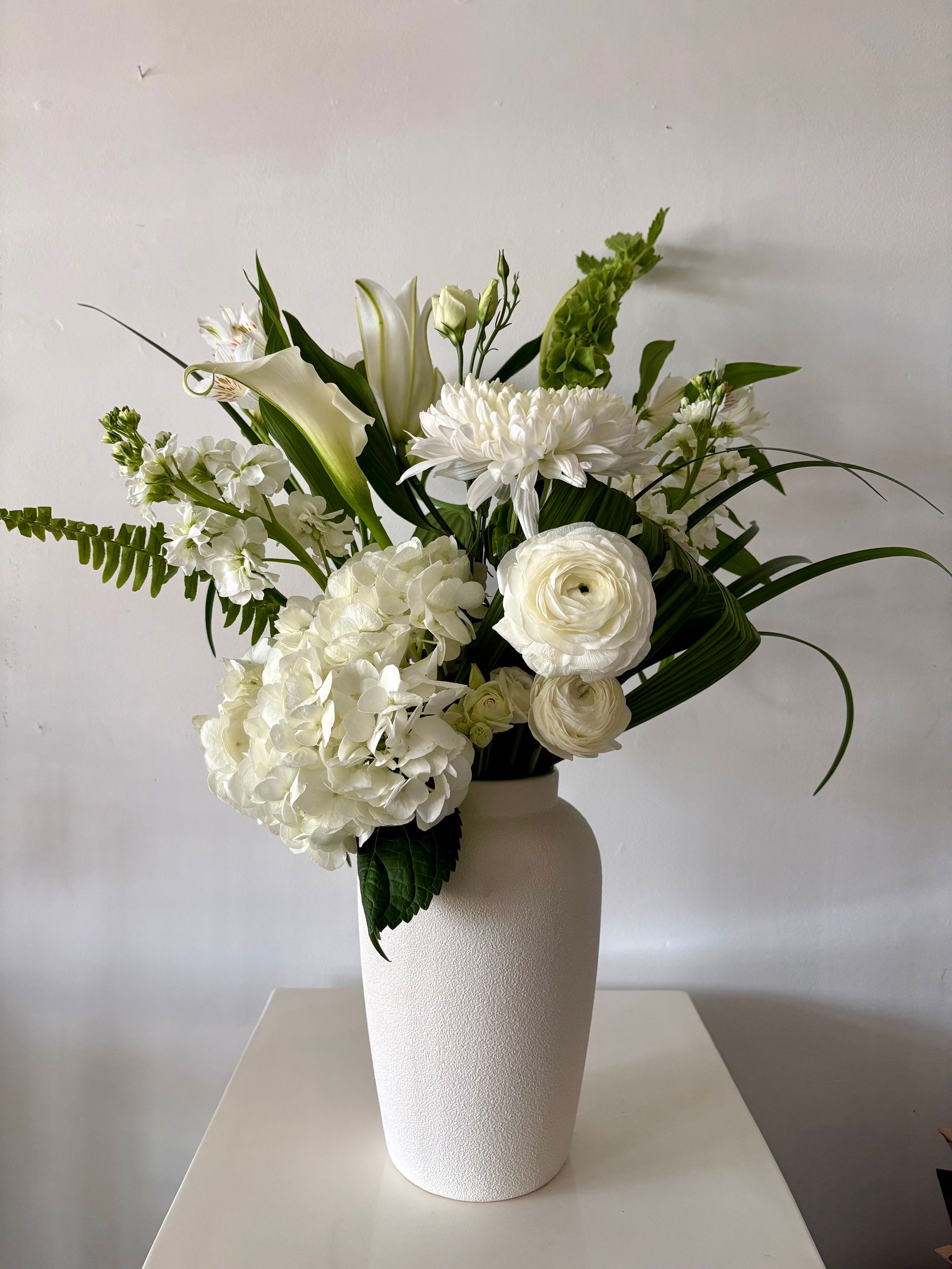 Composition florale blanche dans un vase blanc texturé, posée sur une table devant un mur uni.