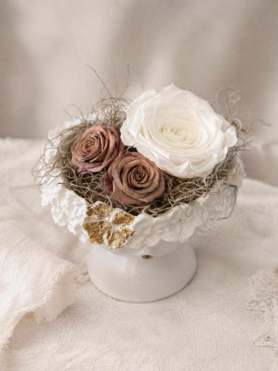 Petit vase blanc avec des roses crème et mauve et des éléments séchés sur un fond en tissu beige clair.