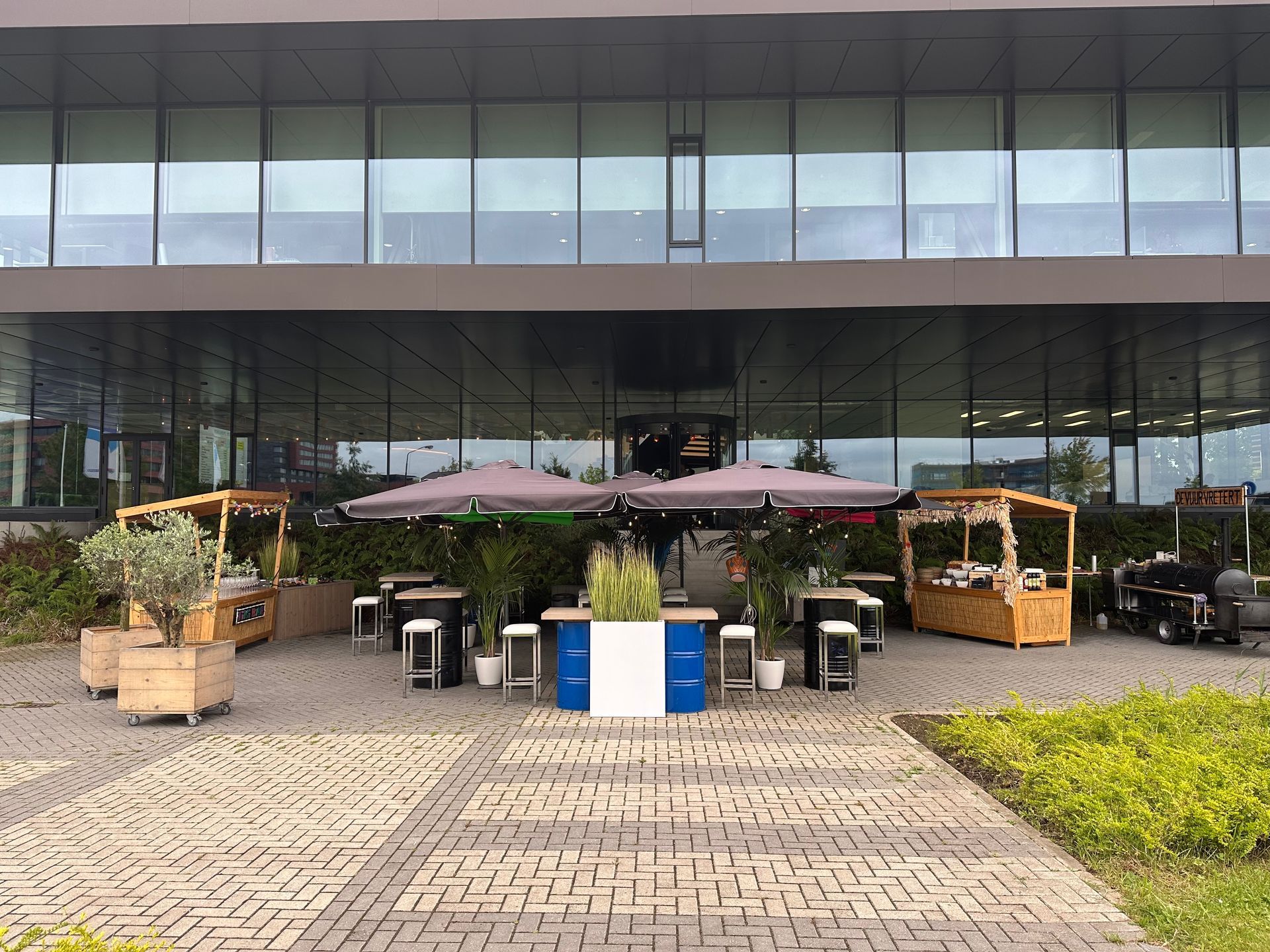 Een terras met tafels en parasols voor een modern gebouw. ​​Er zijn foodtrucks en planten te zien.