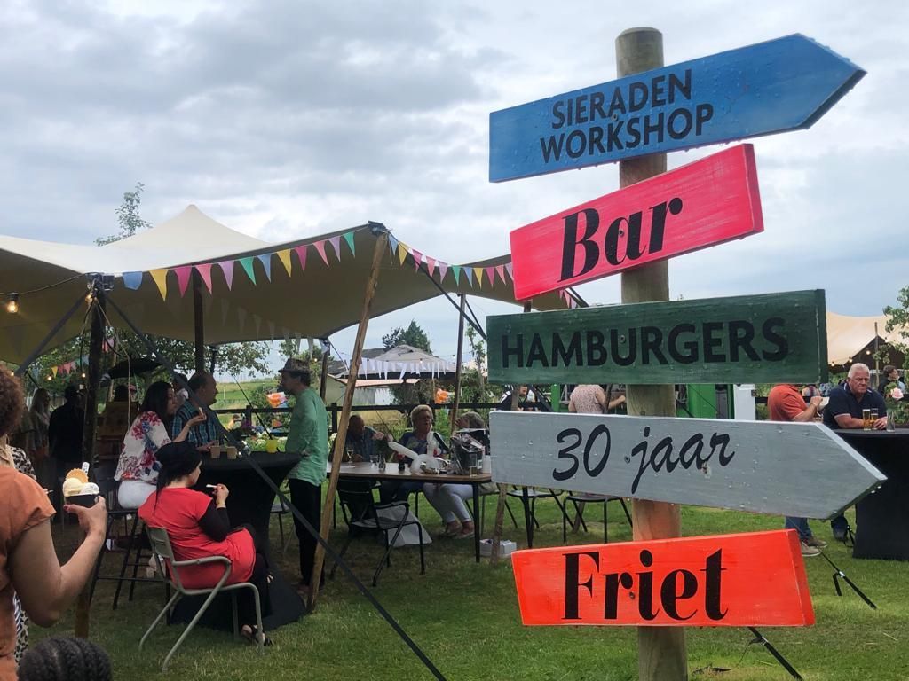 Wegwijzer bij een buitenevenement met routebeschrijving: workshop, bar, hamburgers en friet. Foodtrucks festival