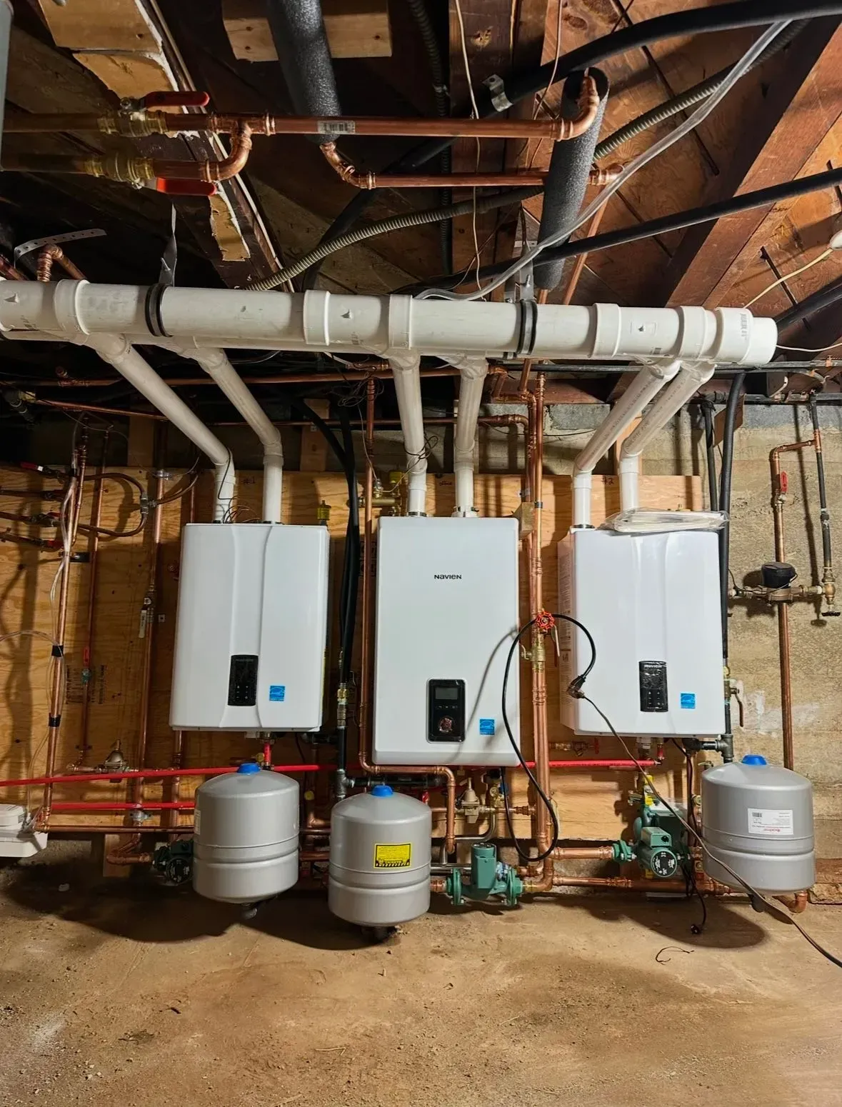 Three white tankless water heaters installed in a basement, connected by pipes and tanks.
