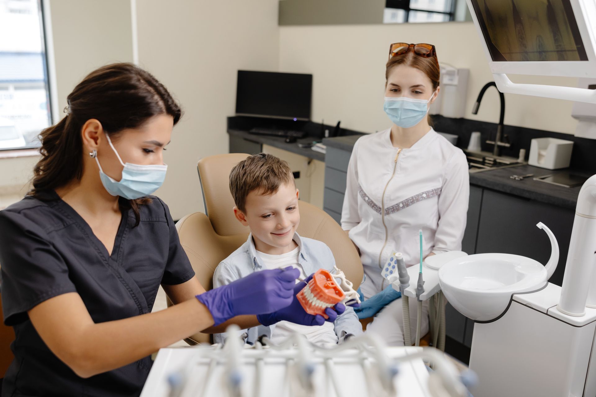 Dentist showing boy a teeth model in clinic; assistant watches.