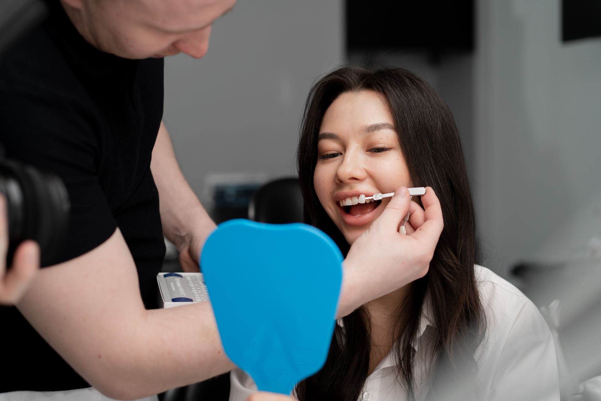 Person examining a woman's teeth with a tool in a dental setting, blue mirror. Woman smiles.