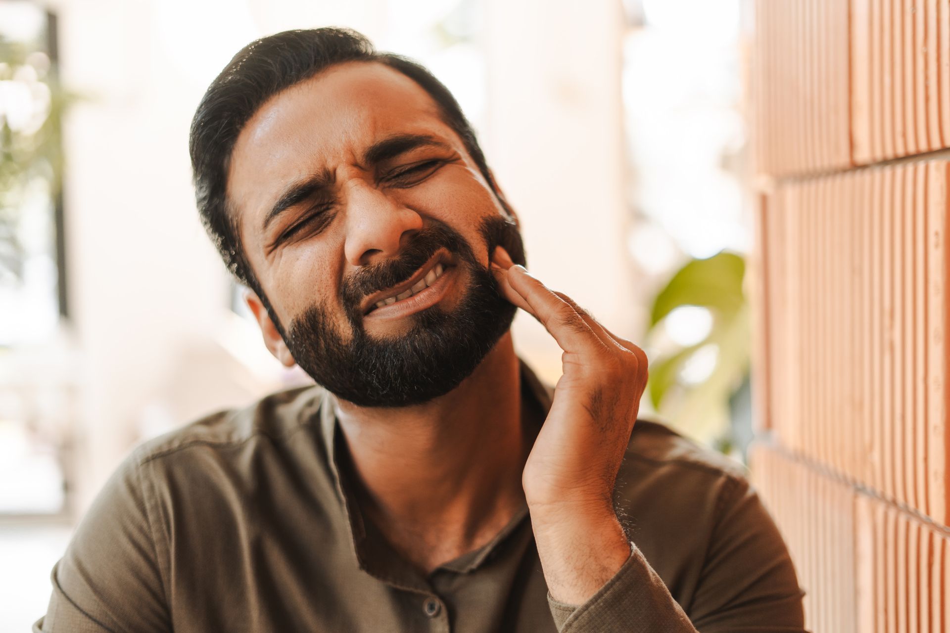 Man with beard grimaces, holding his cheek, appearing in pain.