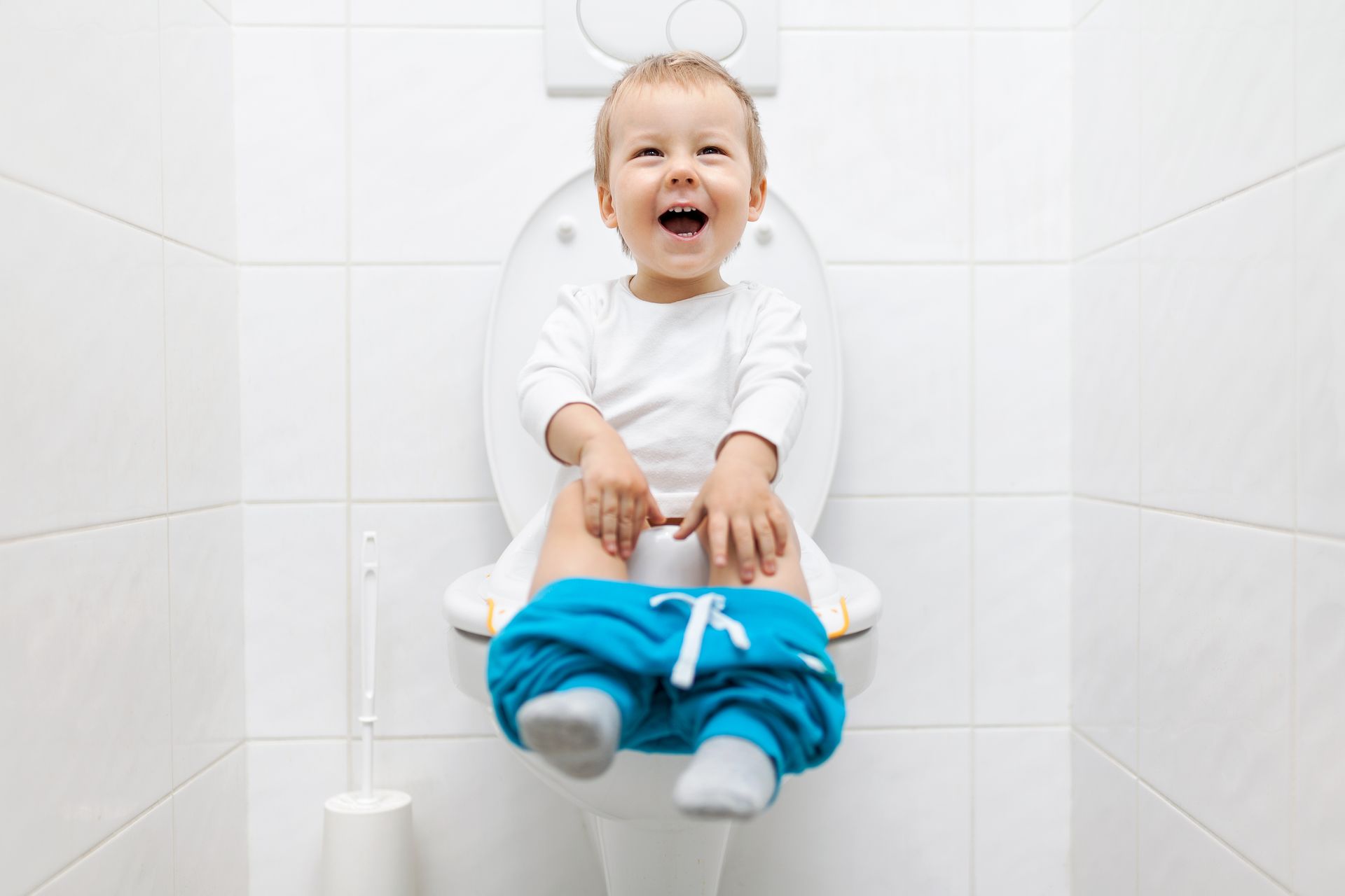 Bebê sorridente sentado no vaso sanitário em um banheiro branco, segurando uma calça azul nos tornozelos.