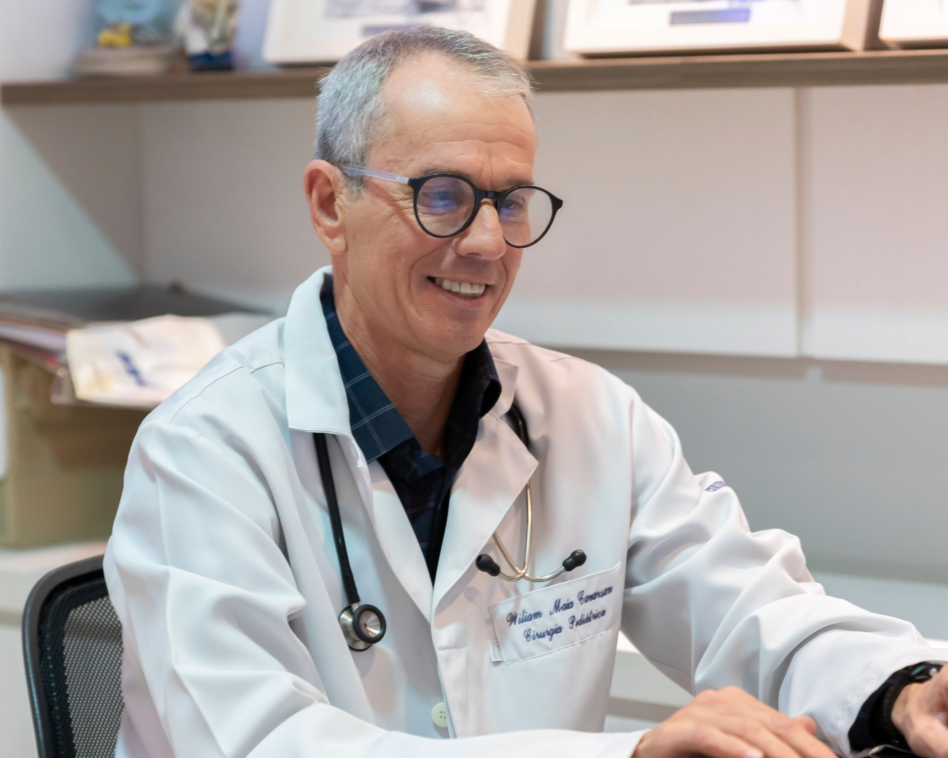 Médico de jaleco branco sorrindo em frente a uma mesa no consultório de uma clínica.