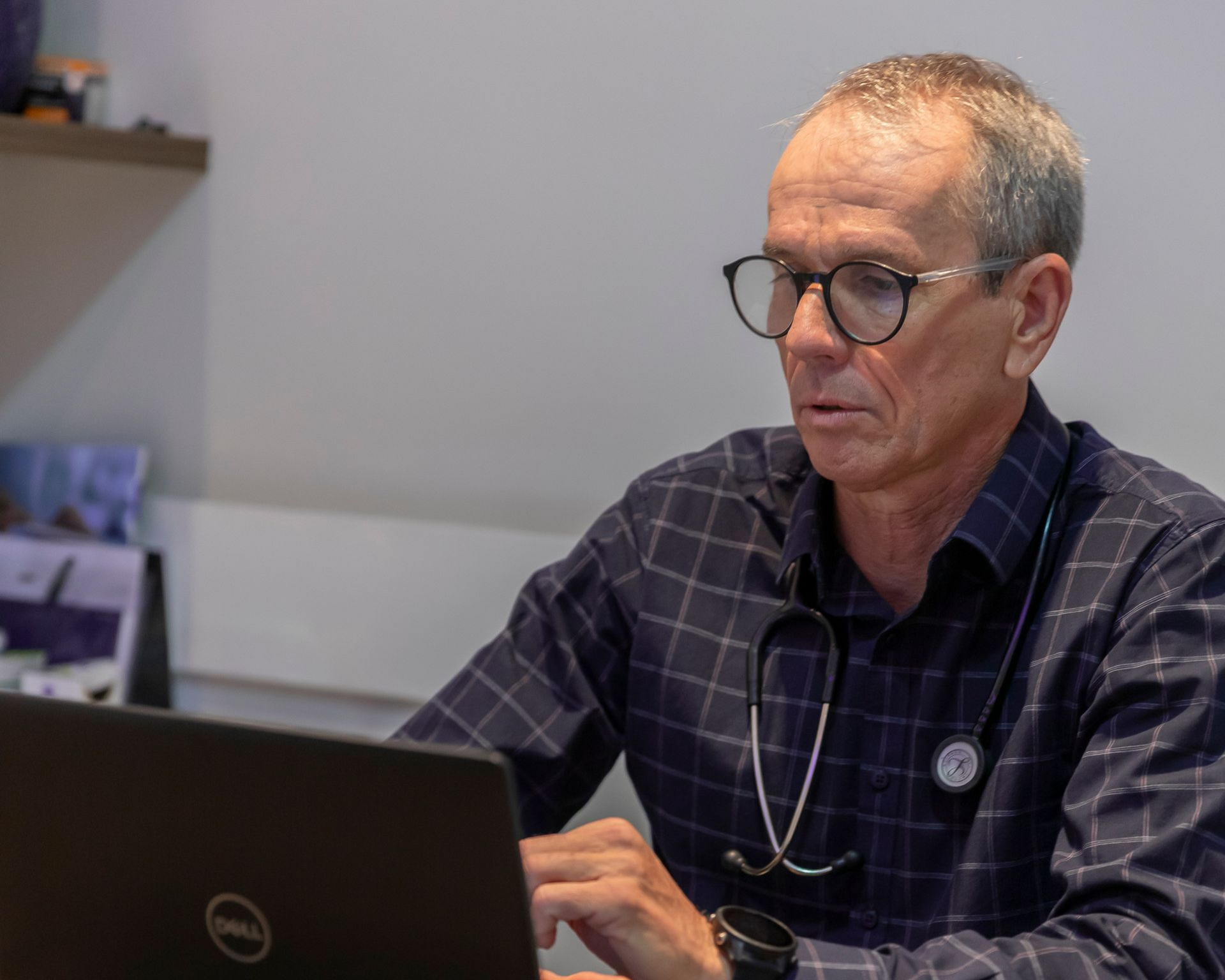 Homem digitando em um laptop em uma mesa, usando óculos e uma camisa xadrez escura.