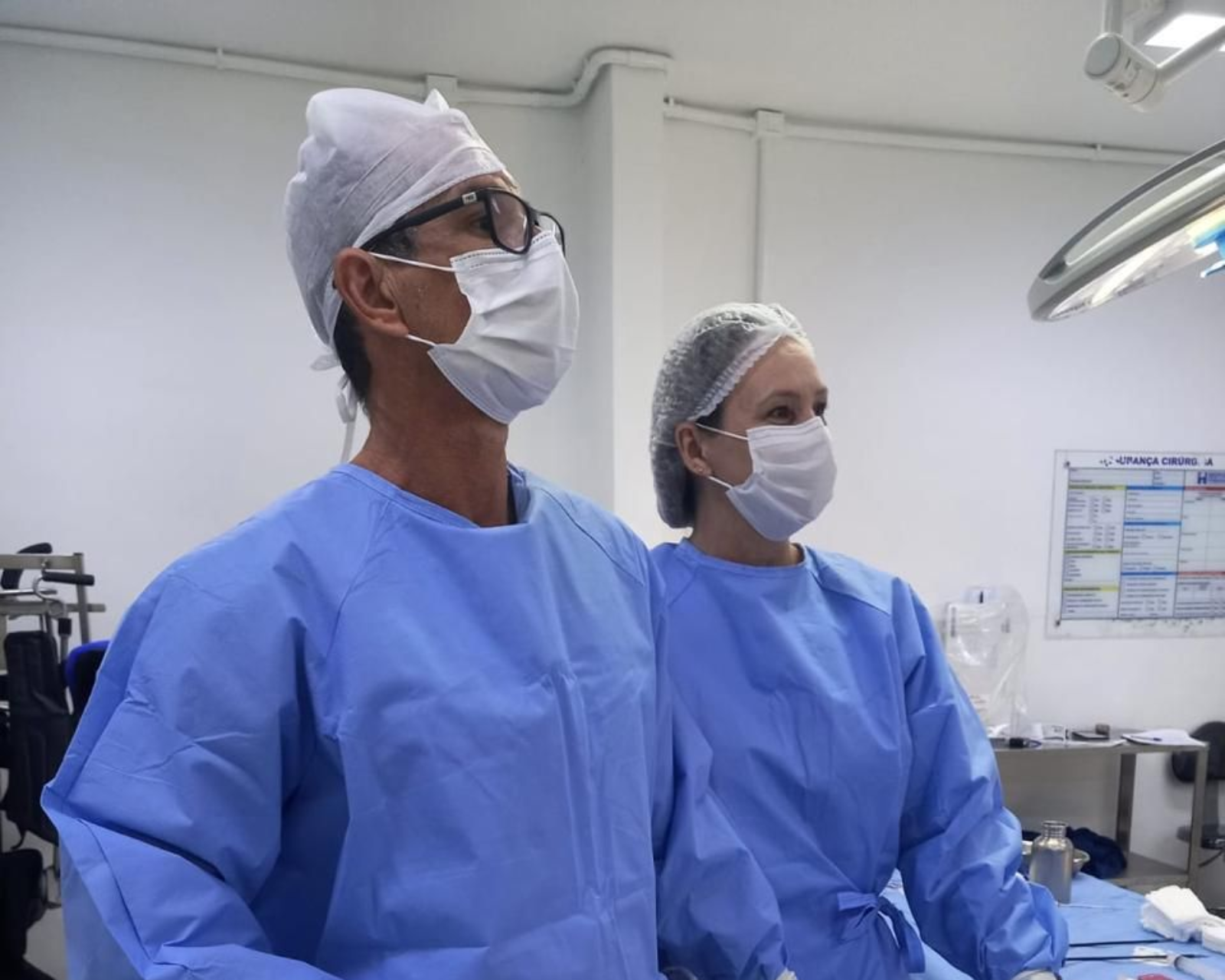 Dois cirurgiões de jaleco azul e máscara em pé numa sala de cirurgia.