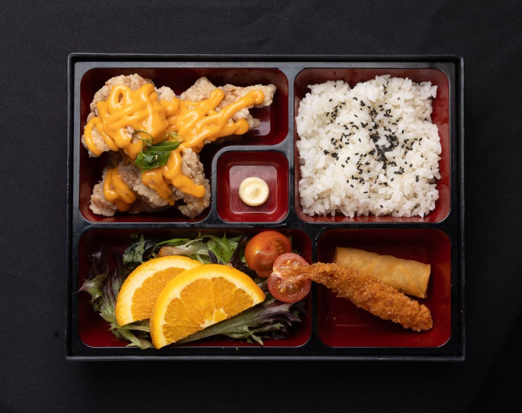 A bento box filled with fried chicken , rice , vegetables and sauce.
