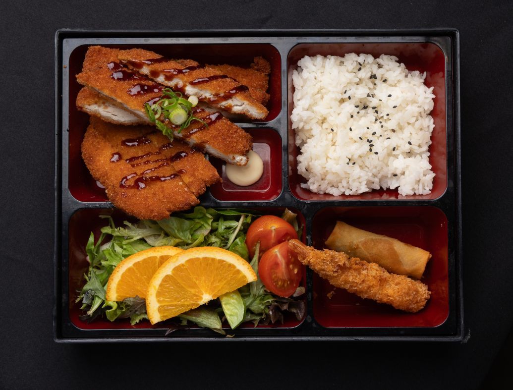 A bento box filled with fried chicken , rice , vegetables , and spring rolls.