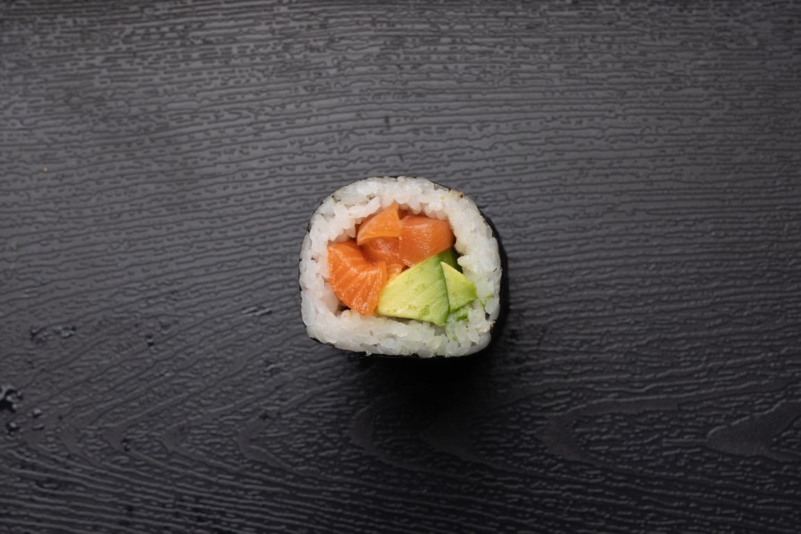 A sushi roll with salmon , avocado and cucumber on a black surface.