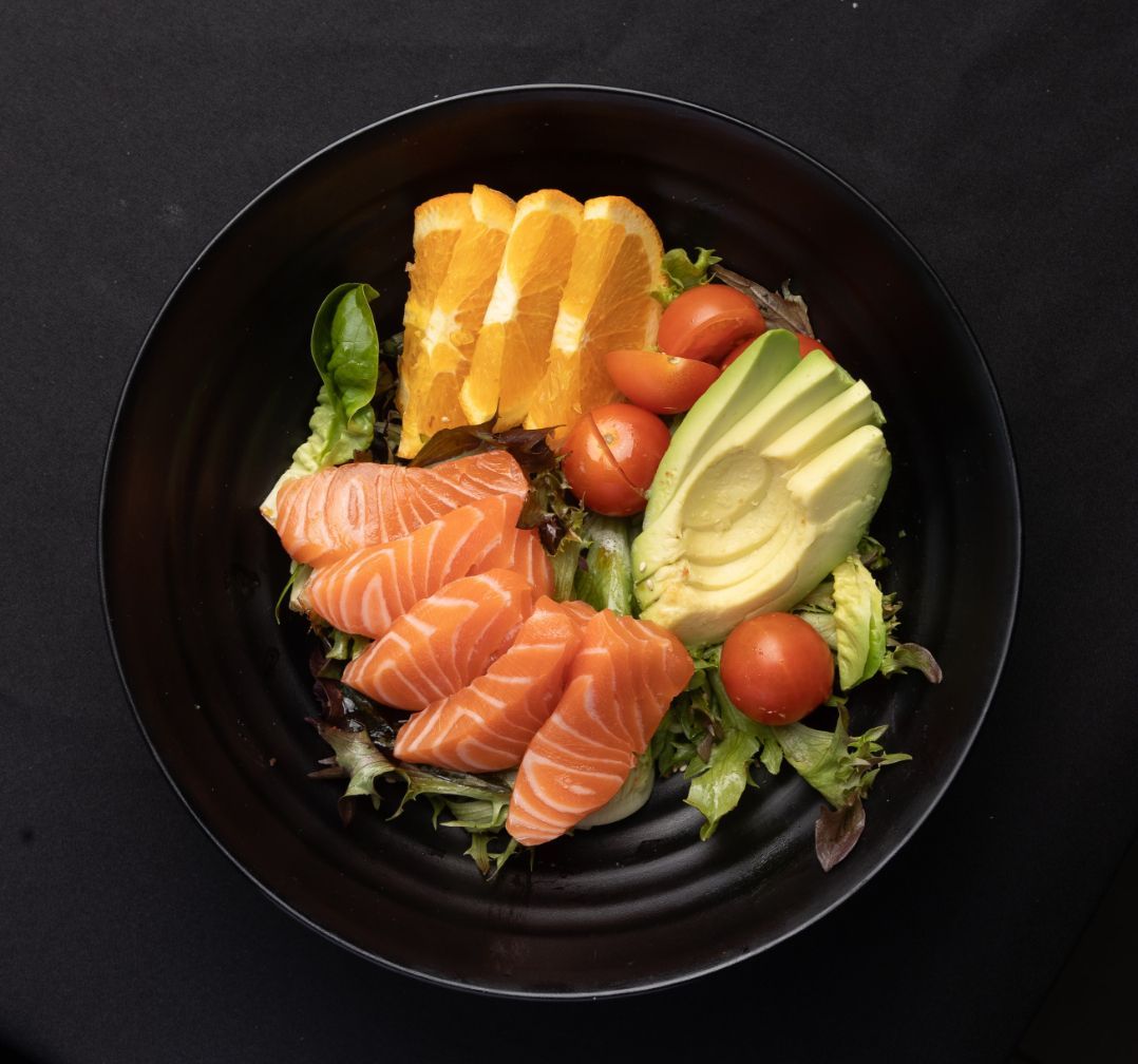 A black bowl filled with salmon , avocado , tomatoes and orange slices.
