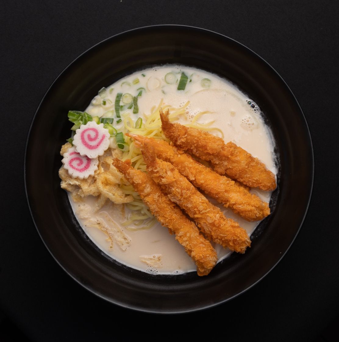 A black bowl filled with noodles and fried chicken on a table.