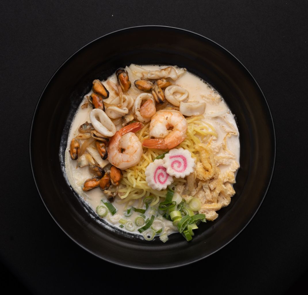 A black bowl filled with noodles , shrimp , mussels and vegetables.