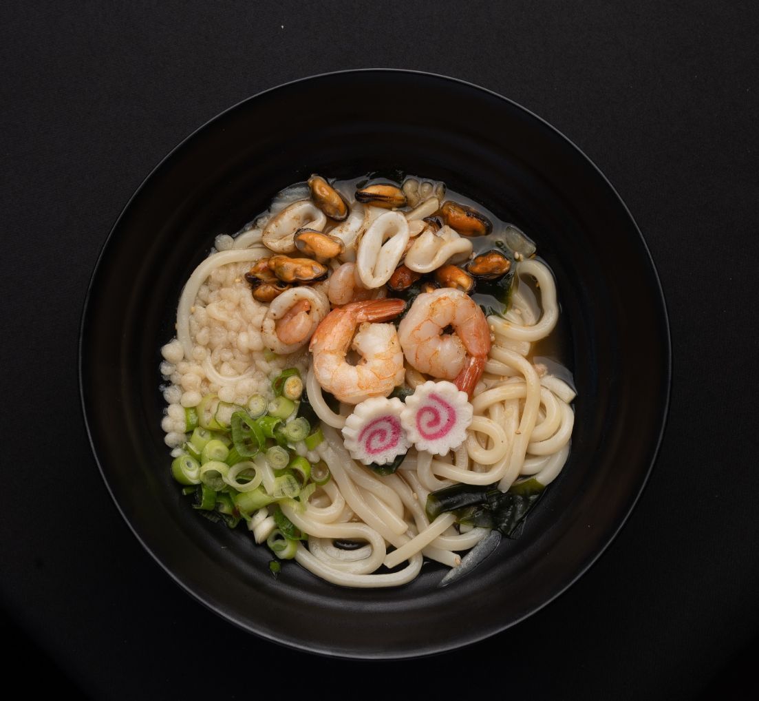 A black bowl filled with noodles , shrimp , squid and green onions.