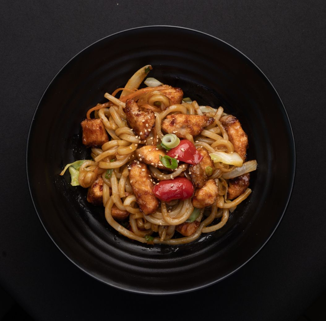 A black bowl filled with noodles and chicken on a black table.