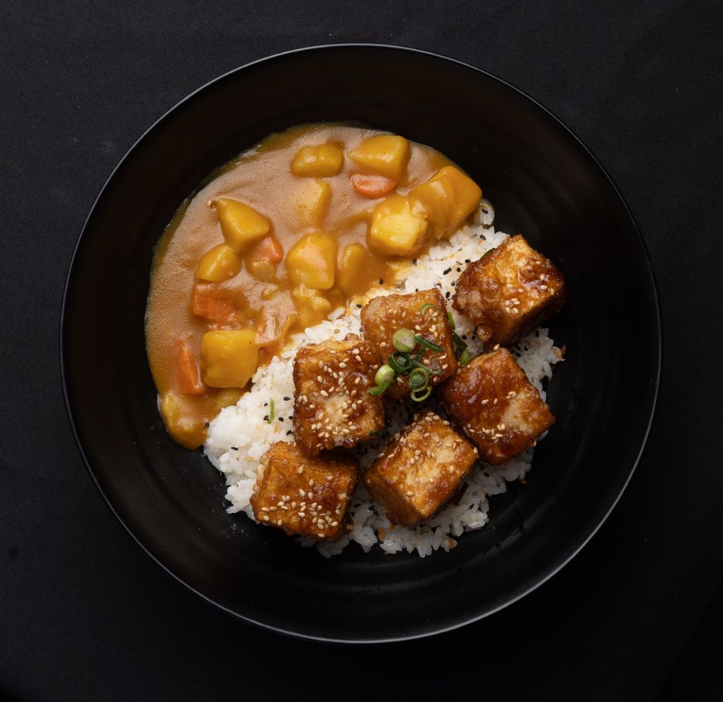 A bowl of curry with rice and tofu on a black table.