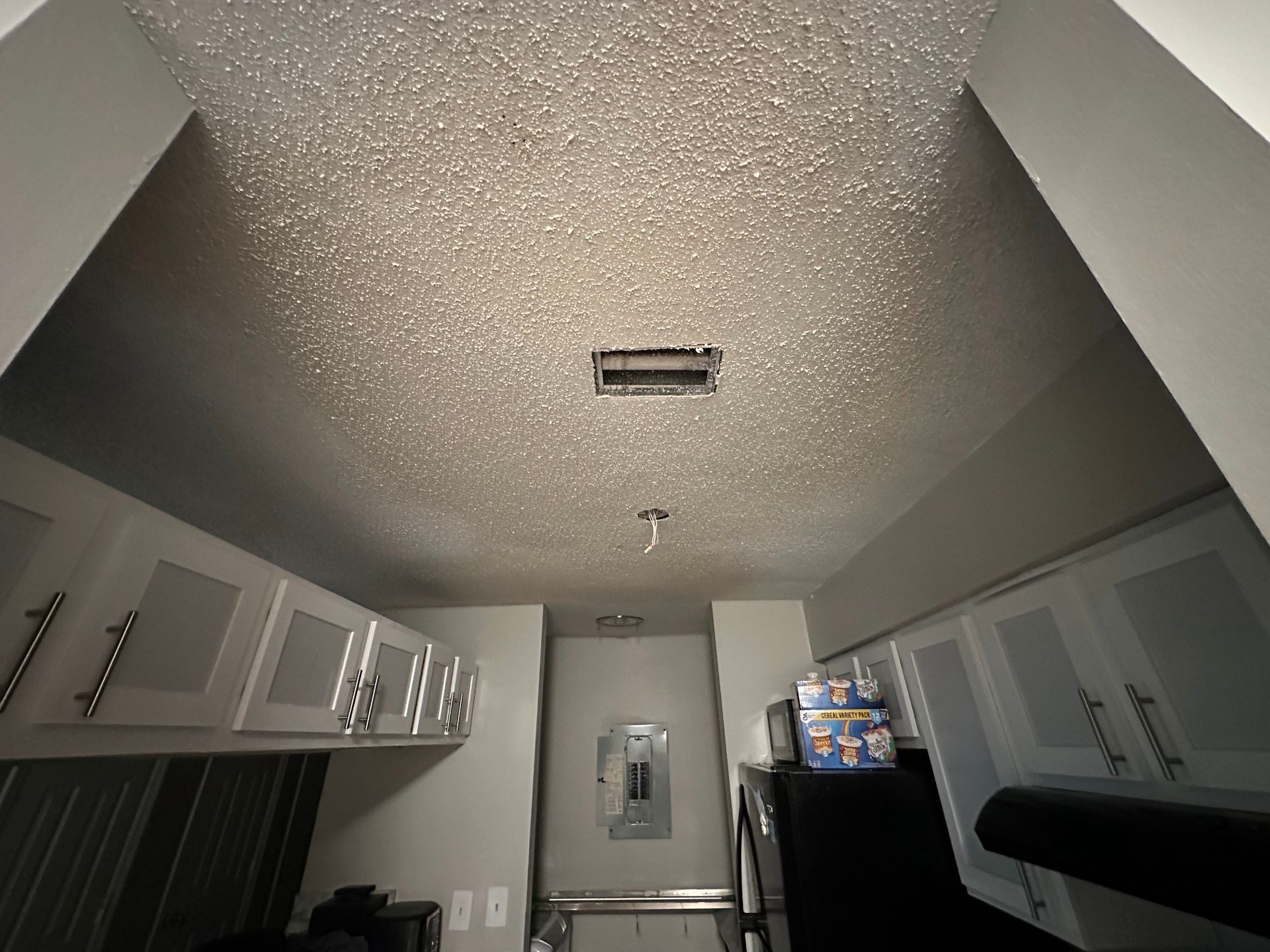 Narrow kitchen with white cabinets, black refrigerator, and textured ceiling under a fluorescent light.