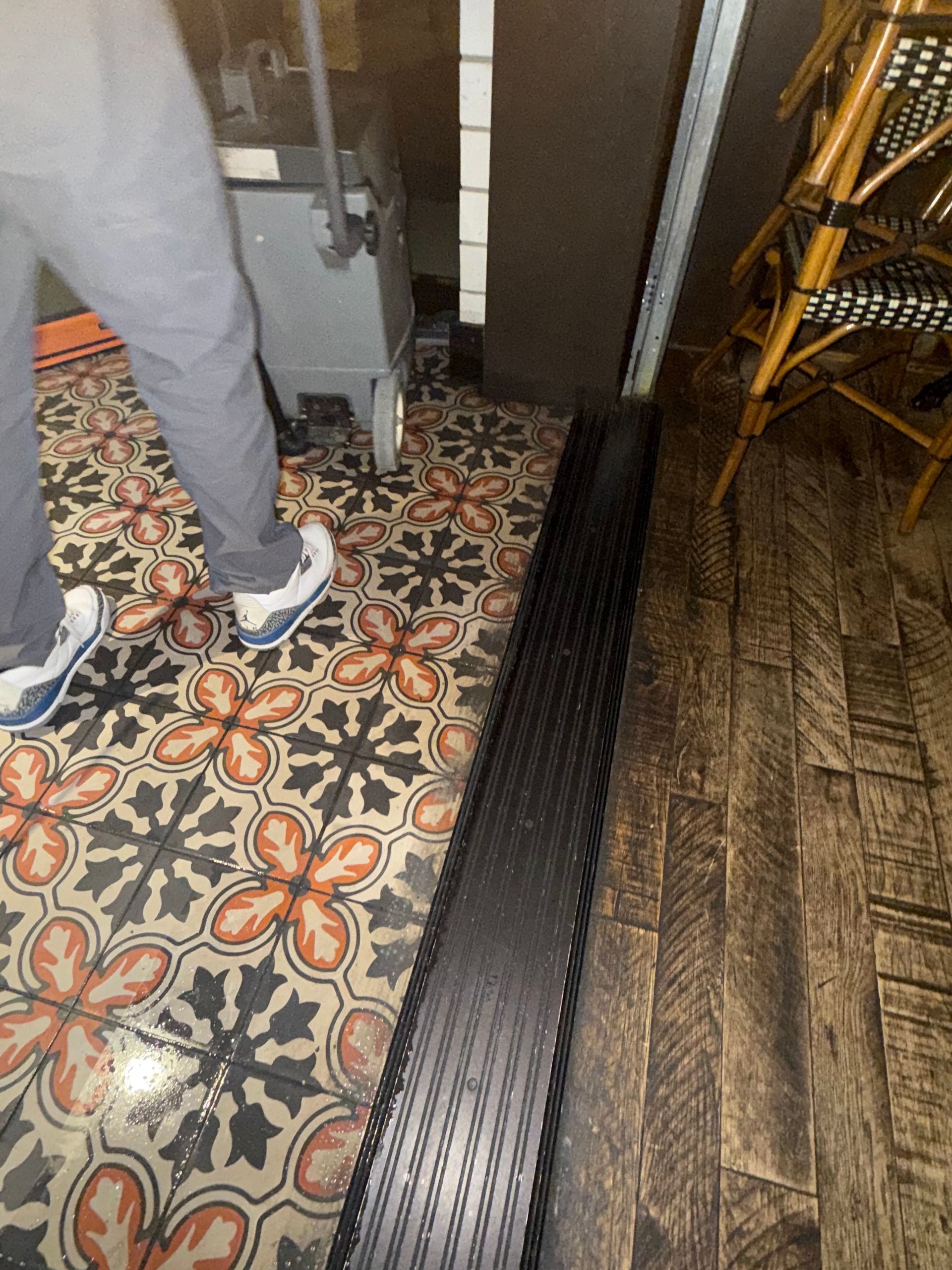 Person pushing a machine over patterned tile, next to a black strip, and wood flooring.