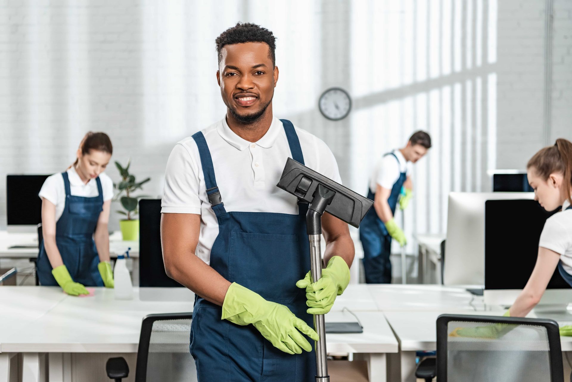 A man in uniform holds a vacuum; cleaning staff works in an office.