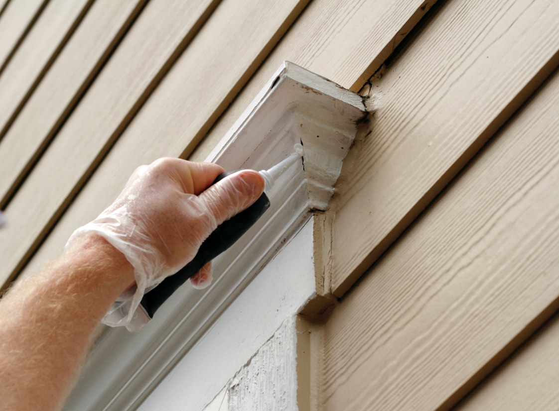 Gloved hand caulk's exterior trim. Beige siding background.