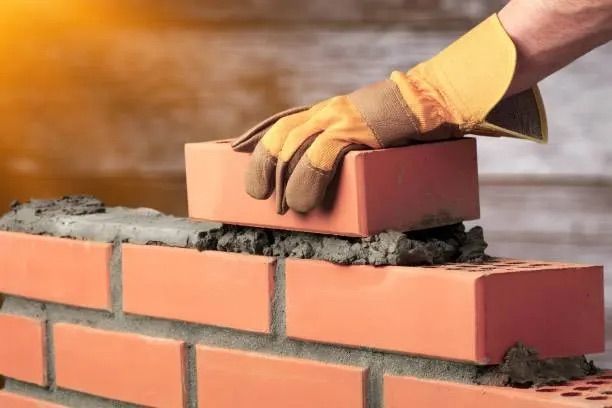 Hand wearing a yellow glove places a red brick on a mortar-covered wall, against a blurred background.