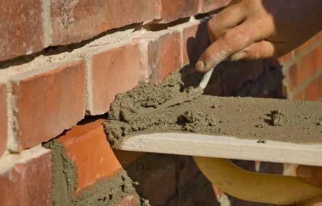 A person using a trowel to apply mortar between red bricks, outdoors.