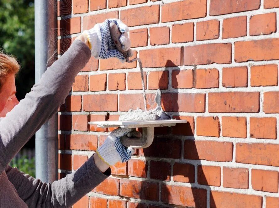 Person wearing gloves applying mortar to a brick wall with a trowel and hawk.