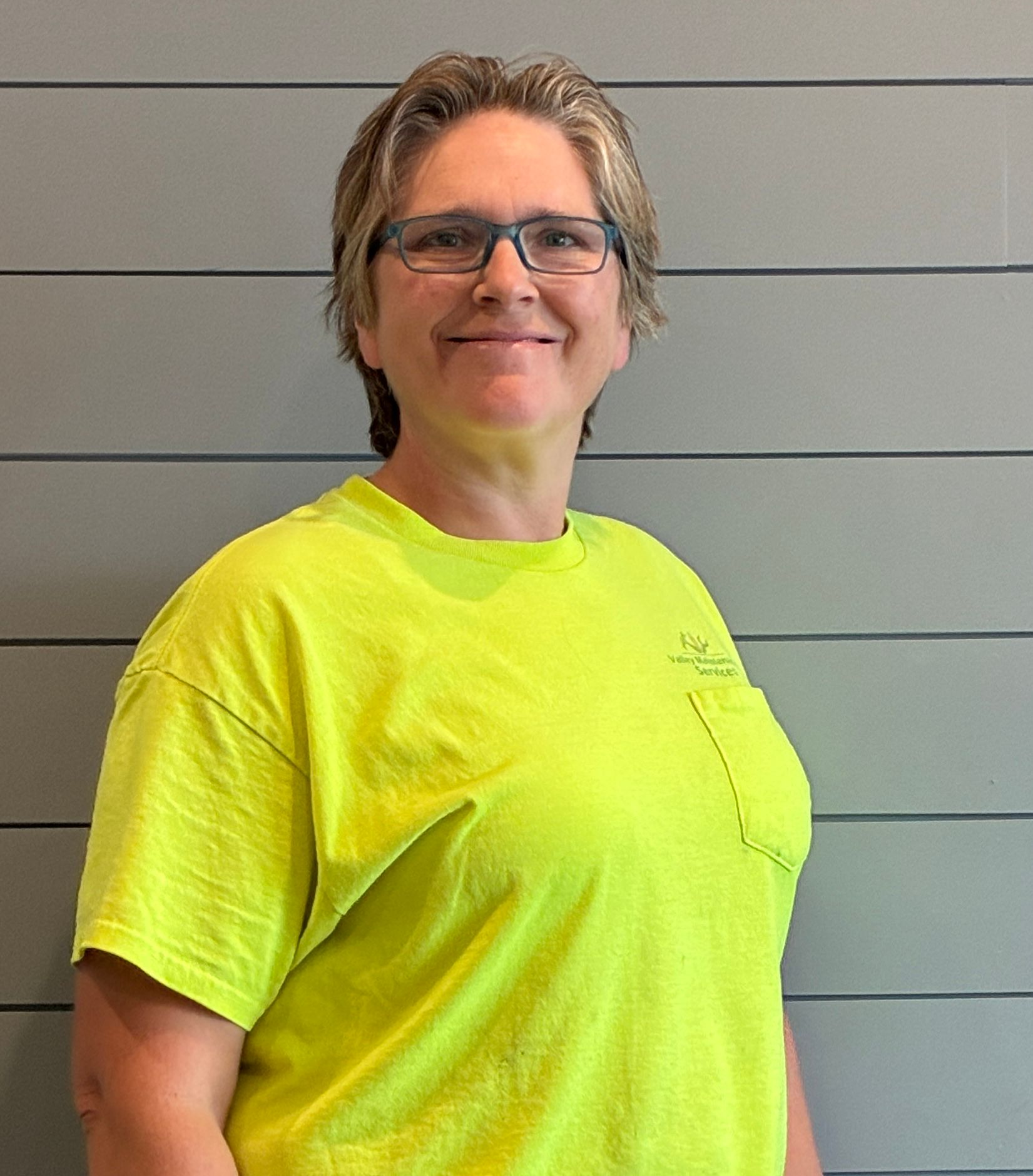 Smiling person wearing glasses and a bright yellow shirt against a gray wall