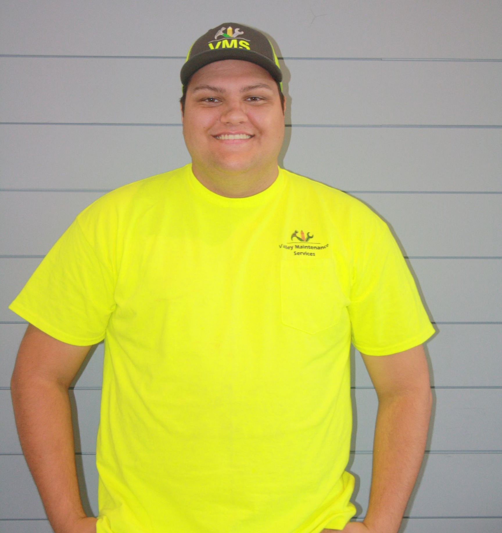 Smiling person in a neon yellow T-shirt and black cap standing against a gray wall