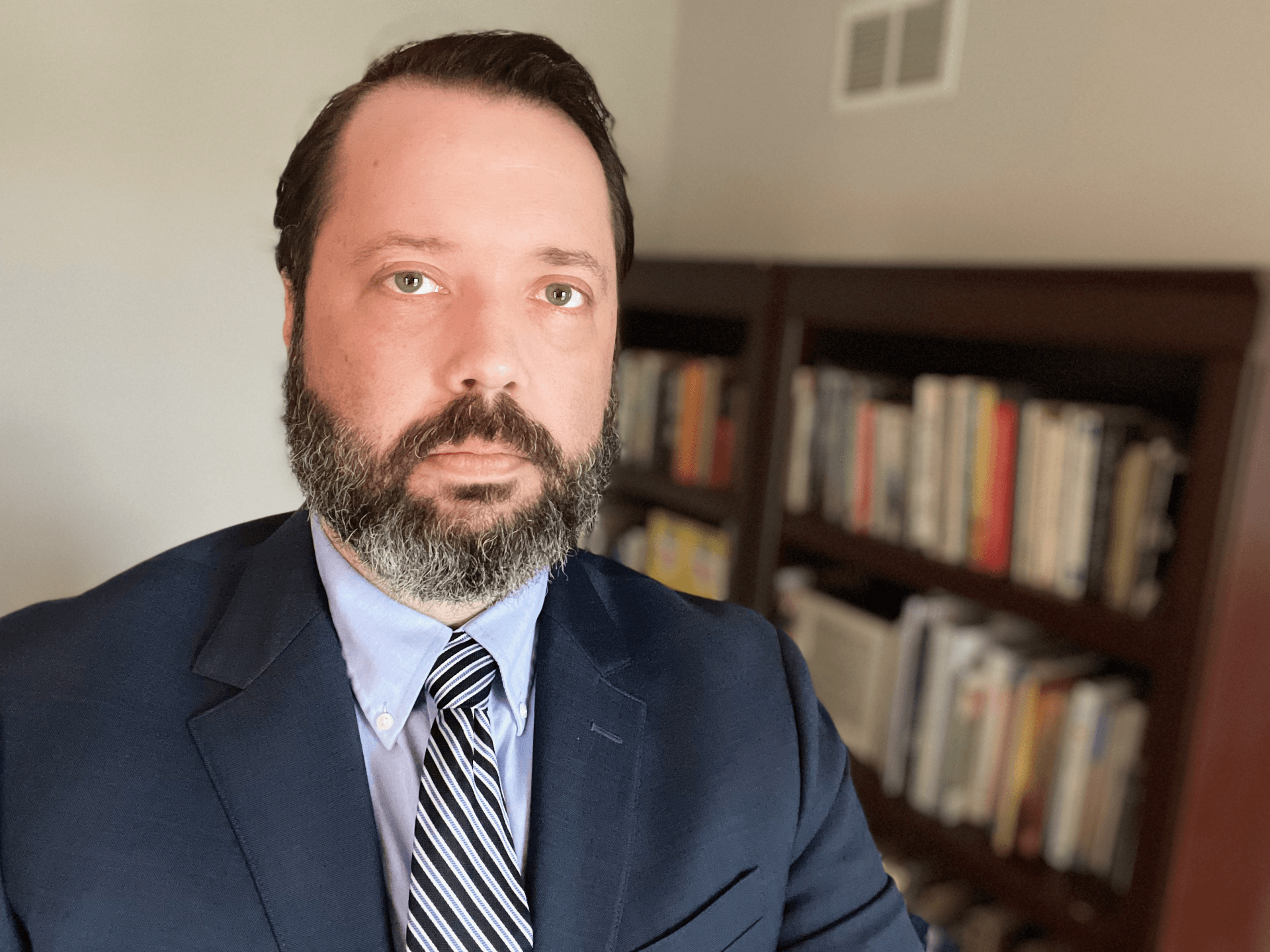 Man with a beard in a suit and tie, looking at the camera; bookshelves in the background.