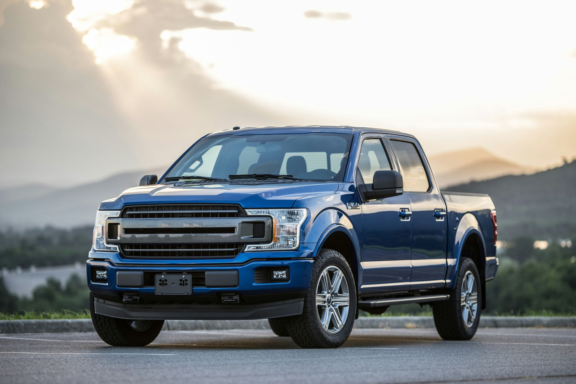 Blue Ford F-150 pickup truck parked on asphalt road with mountains and sun in the background.